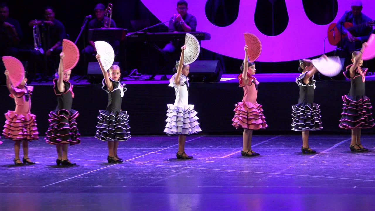 A row of young dancers in colorful dresses raise folding fans overhead on a purple-lit performance stage in Cabo San Lucas, Mexico.