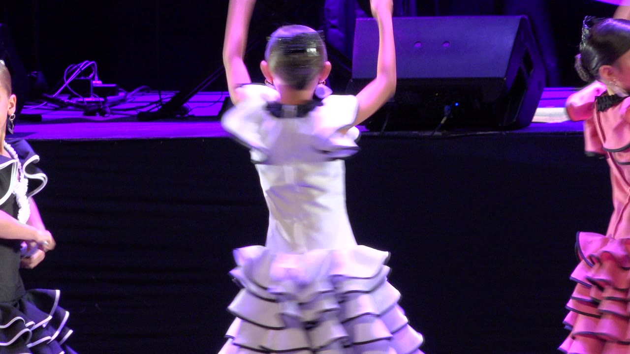 A girl in a white ruffled dress is seen from behind with both arms raised while another girl in pink turns beside her on the Cabo San Lucas stage.