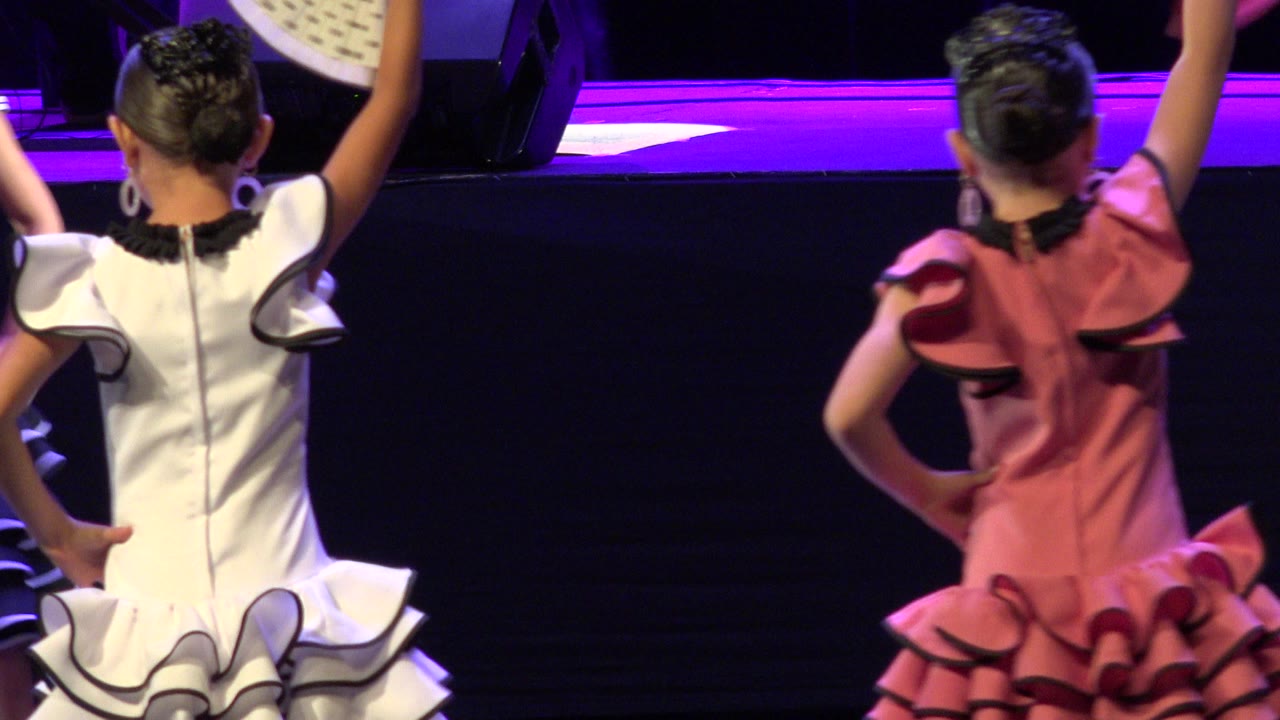 Two girls seen from behind in ruffled stage dresses with one arm raised.