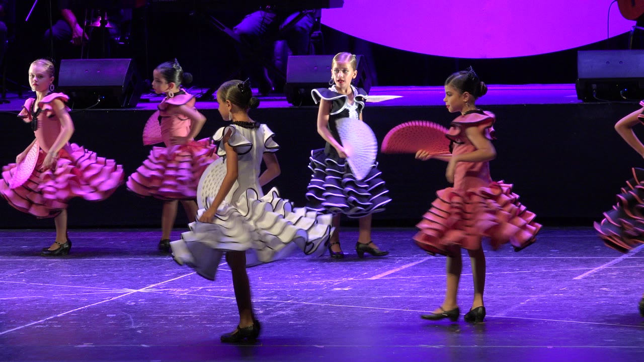 Girls in flamenco-style dresses dance with fans on a purple-lit stage.