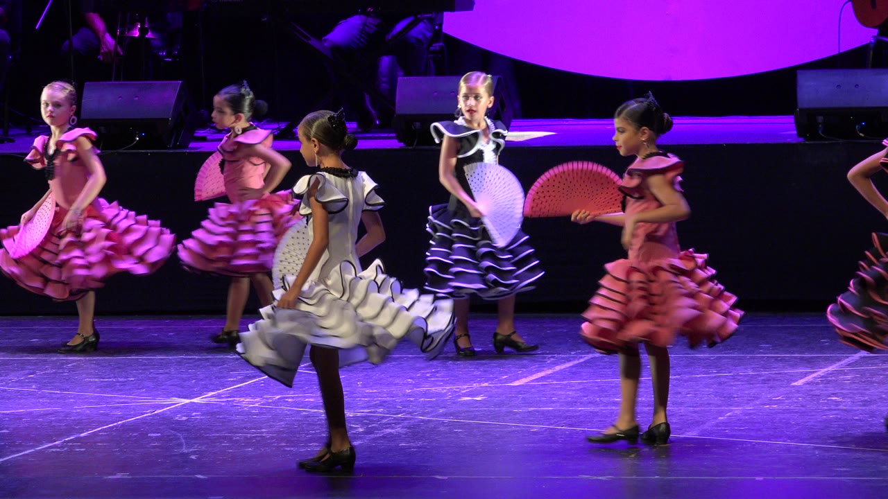 At 0:01 in Cabo San Lucas, Mexico, several young dancers in pink and white dresses pass each other across the stage while a girl in black holds a white fan near center.