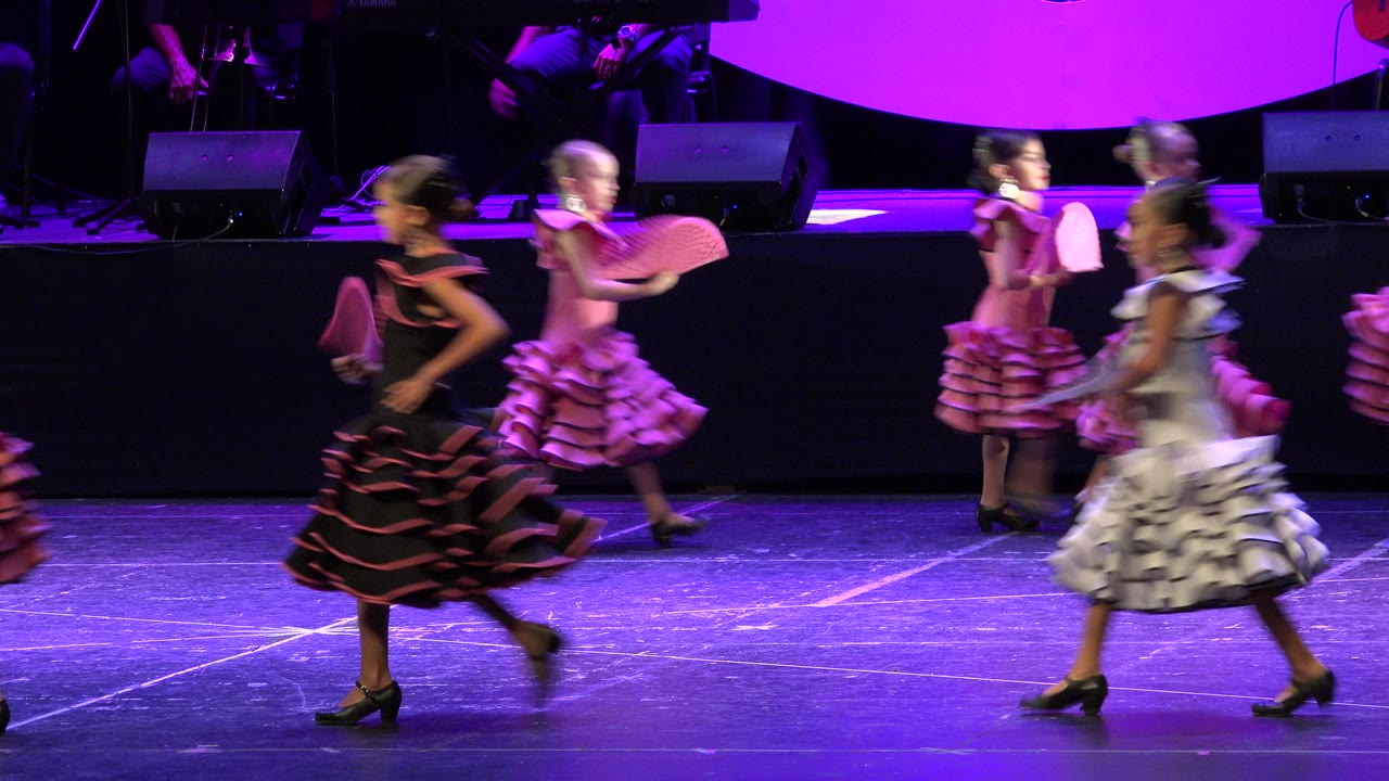 Young girls in ruffled dresses perform with folding fans on a purple-lit stage in Cabo San Lucas, Mexico, with musicians partly visible in the background.