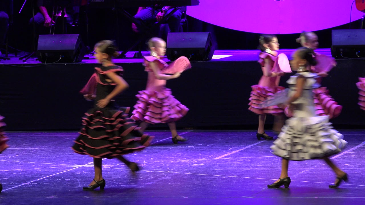 Several young dancers in pink, black, and white Spanish-style dresses move in opposite directions under purple stage light in Cabo San Lucas, Mexico.