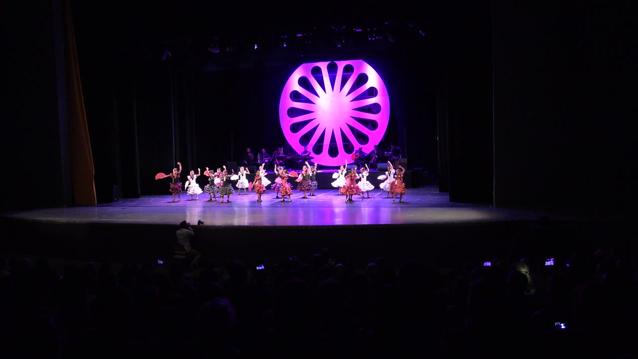Wide audience-view frame in Cabo San Lucas showing performers spread across a lit theater stage beneath a glowing pink-purple circular backdrop.