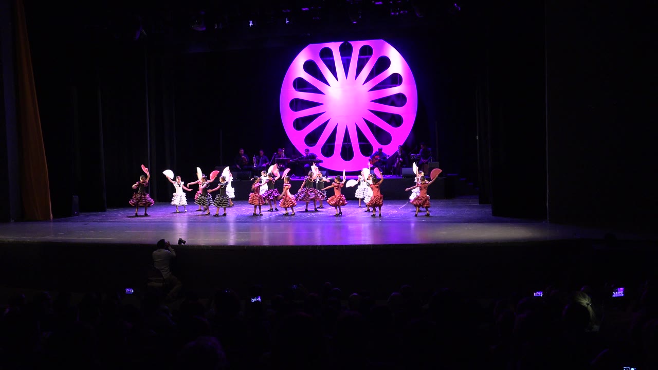 A wide auditorium view in Cabo San Lucas, Mexico shows dancers spread across a stage beneath a large glowing magenta circular backdrop at the start of the routine.