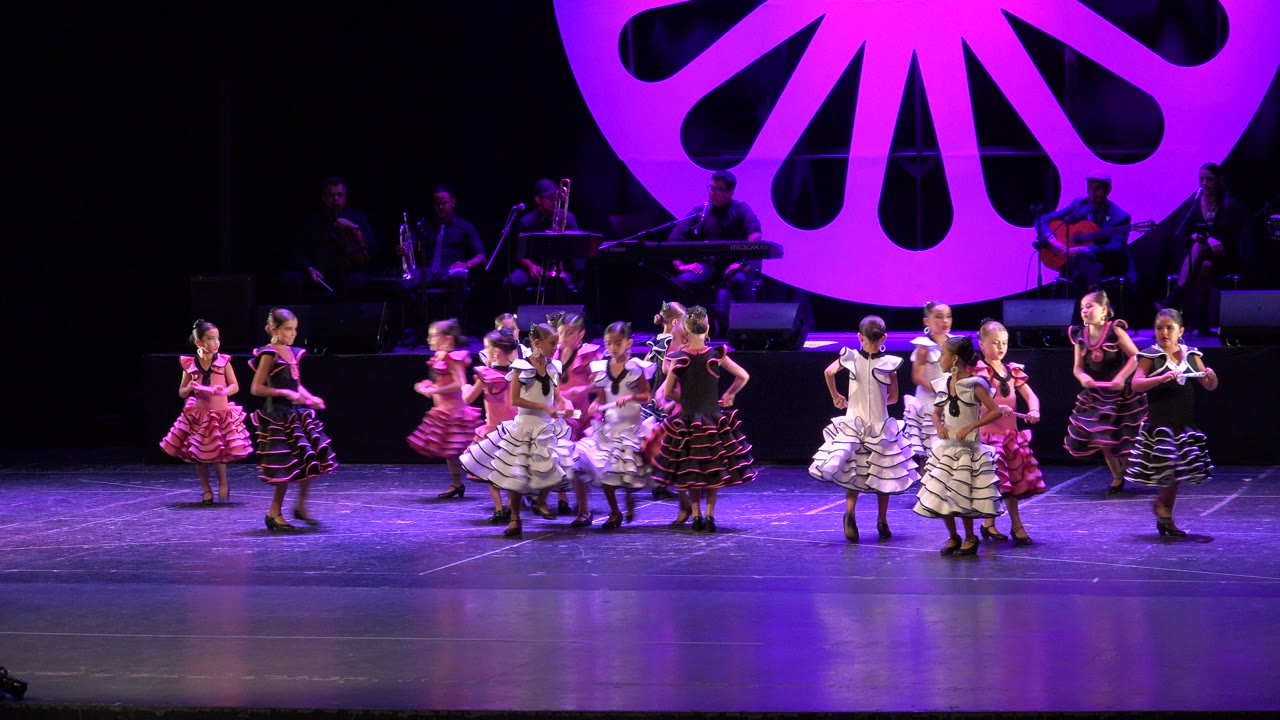 Children in white, pink, and black ruffled dresses move through the center of the stage in Cabo San Lucas, Mexico, while the band remains in shadow behind them.