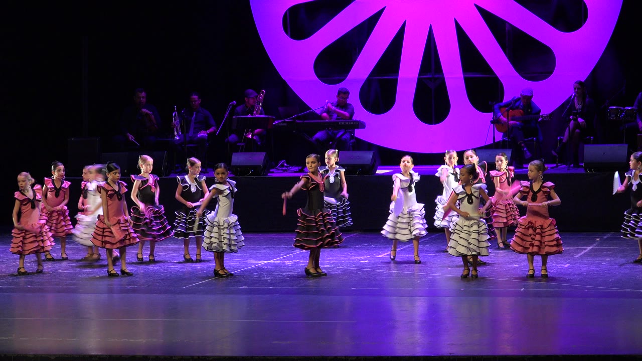 Dancers in pink, black, and white ruffled dresses perform on a purple-lit stage in Cabo San Lucas, Mexico, with a large shell-shaped set piece behind them.