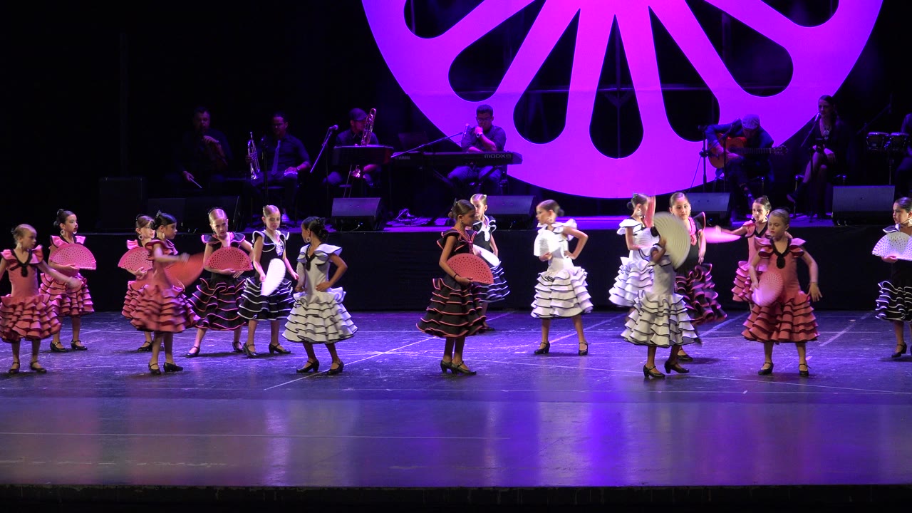 Young dancers in ruffled dresses perform with fans on a purple-lit stage.