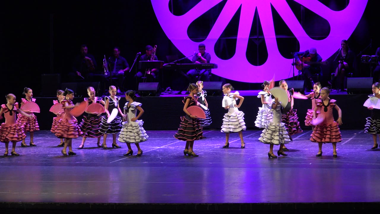 At 0:01, the dancers continue their stage routine in Cabo San Lucas with fans held at chest and shoulder height, framed by purple light and a shadowed live band.