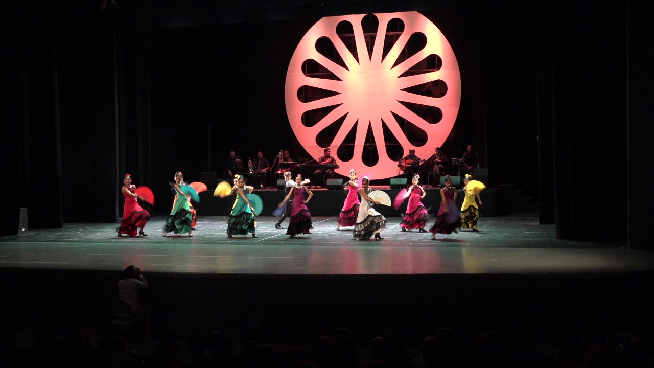 Wide Cabo San Lucas performance frame with dancers shifting positions across the stage while the illuminated radial backdrop and seated musicians remain in the background.