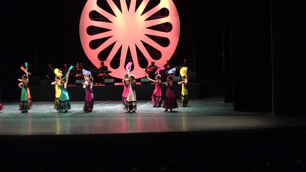 A wide view in Cabo San Lucas, Mexico shows young performers holding fans at different heights while the band sits in shadow behind a glowing pink set piece.