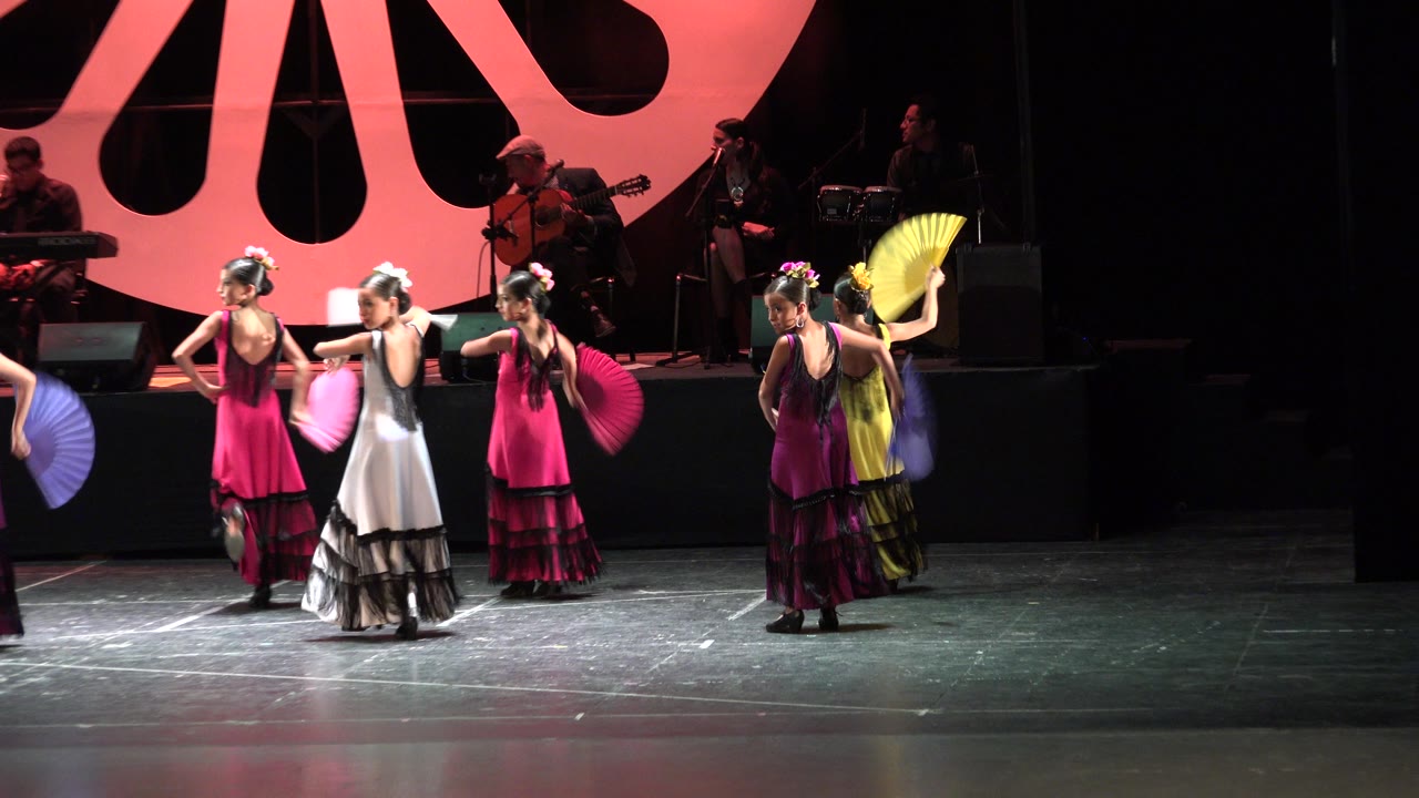 Young girls perform with folding fans on a dark stage in Cabo San Lucas, Mexico, with musicians visible behind them under red lighting.