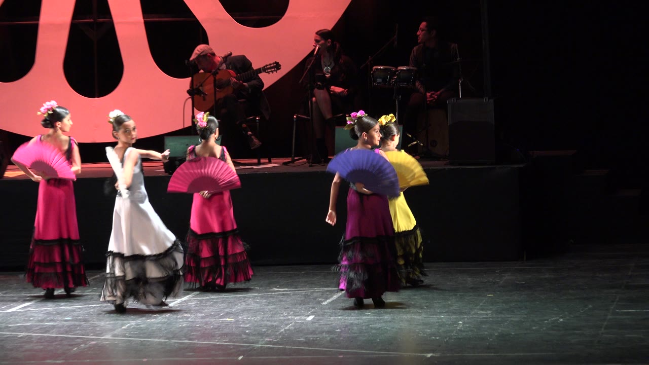 Girls turn across the stage with fans and ruffled dresses.
