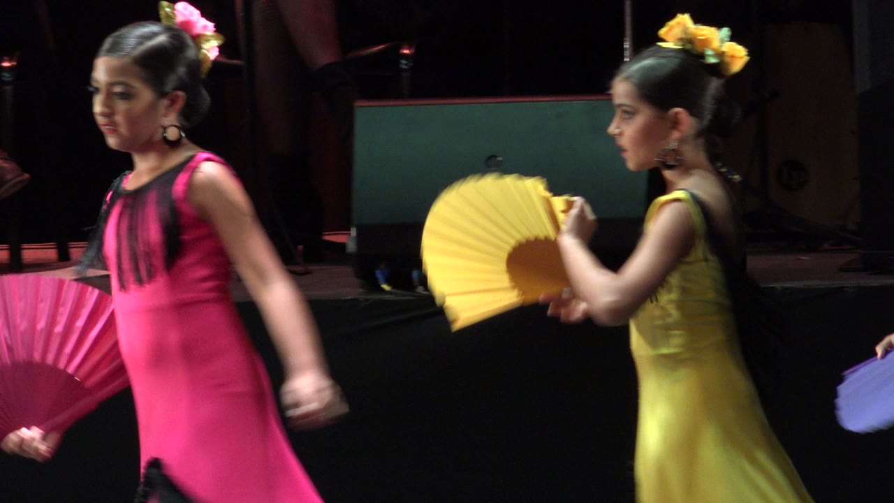 Two young girls perform with pink and yellow folding fans on a dark stage in Cabo San Lucas, Mexico, with stage equipment behind them.