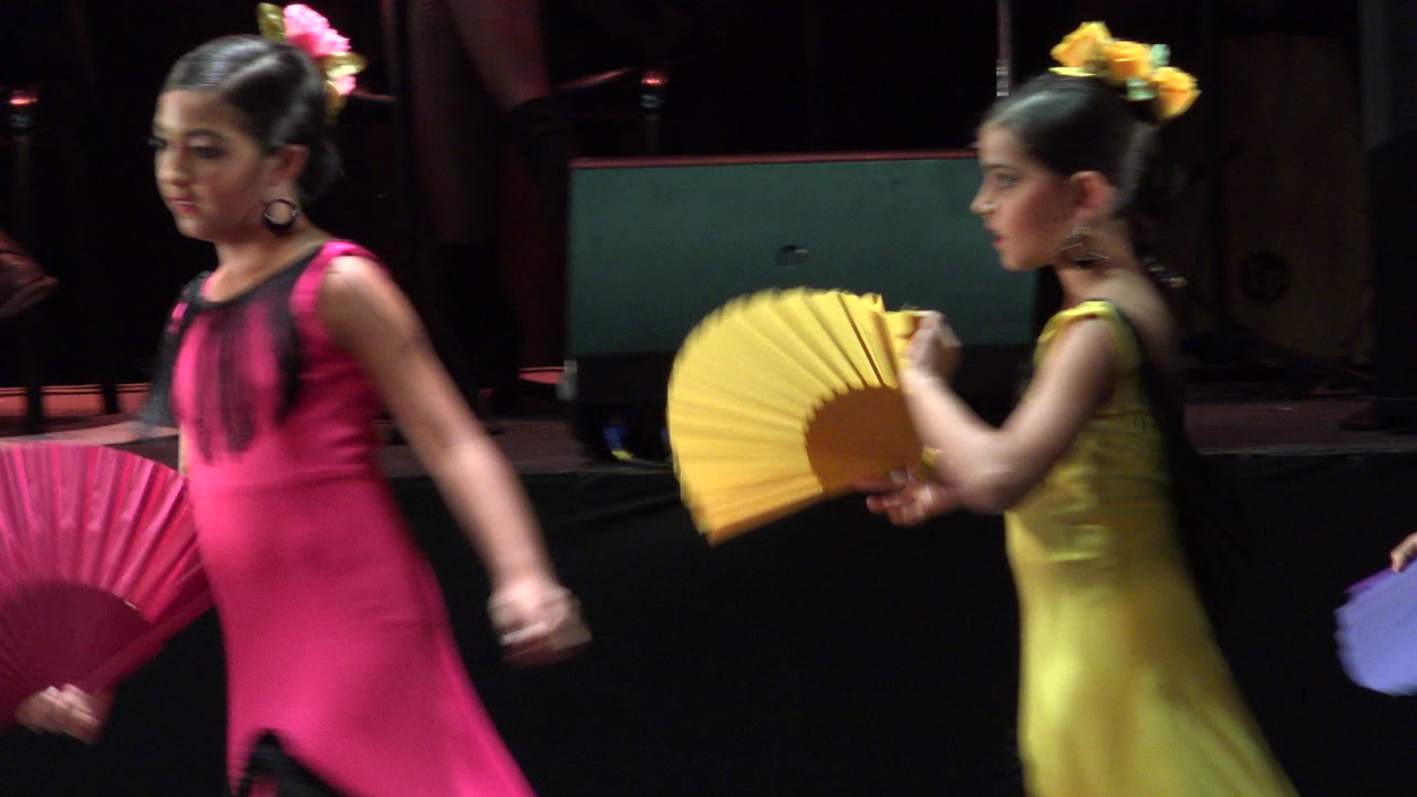 A young girl in a pink dress moves across the foreground while another young girl in yellow holds a yellow fan on the Cabo San Lucas stage.