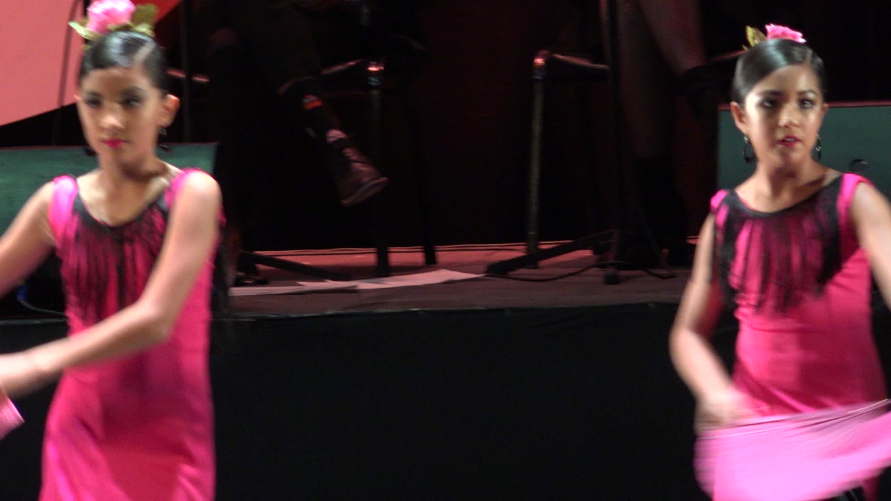 At 0:01, two performers in matching pink dresses appear side by side on the Cabo San Lucas stage as a fan sweeps through the foreground.