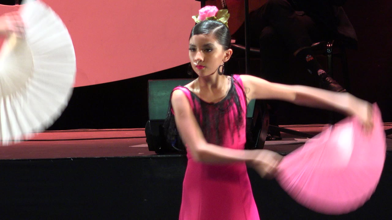 At 0:01, the pink-costumed young dancer faces forward with one arm extended as a blurred pink fan sweeps across the stage in Cabo San Lucas, Mexico.