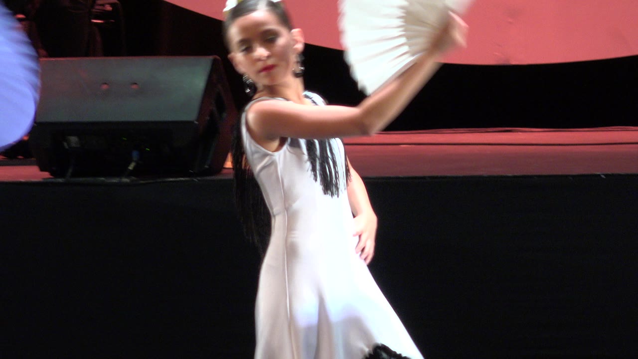 The white-dressed child dancer turns her body while lifting a white fan, with the dark stage front and background speaker still visible in Cabo San Lucas, Mexico.