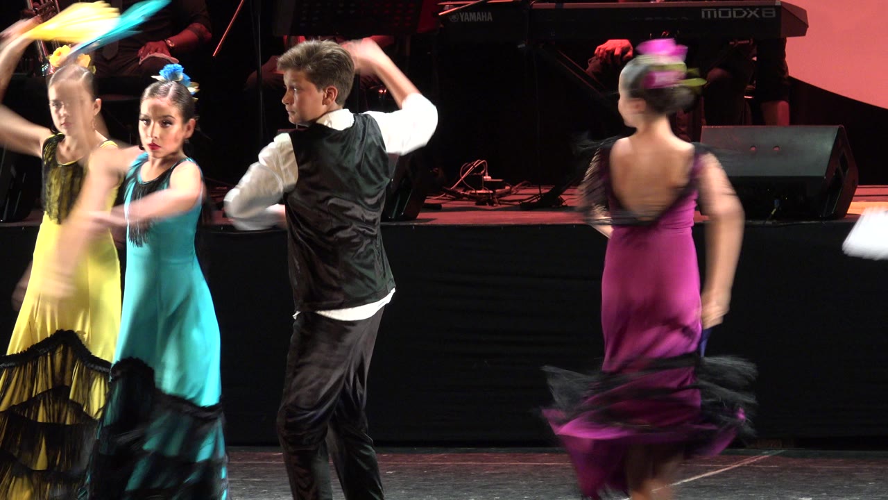 Children perform a choreographed dance on a stage in Cabo San Lucas, Mexico, with a boy in a black vest and girls in teal, yellow, and magenta dresses moving fans under warm stage light.
