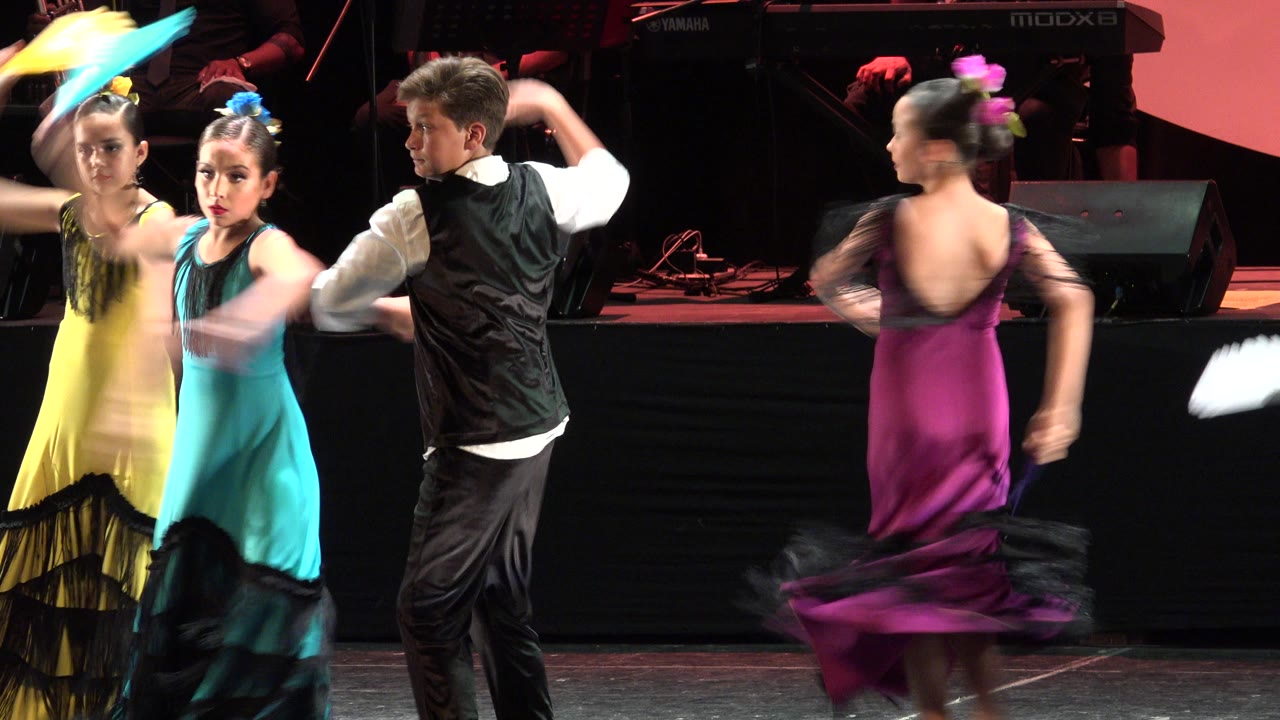 The Cabo San Lucas stage performance continues as two girls in yellow and teal dresses face forward, a boy in a black vest turns sideways, and a magenta-dressed dancer blurs in motion.