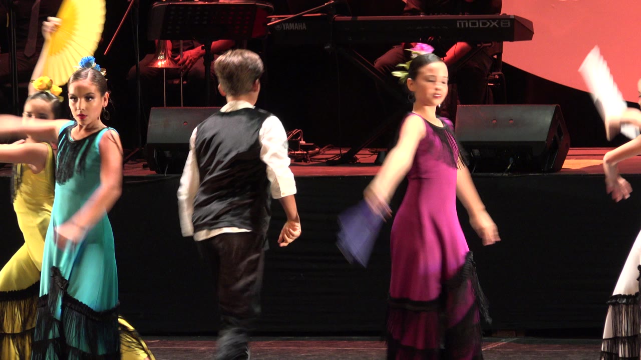 A boy in a black vest and girls in bright dresses move across the Cabo San Lucas stage as a magenta-costumed dancer turns near the right side.