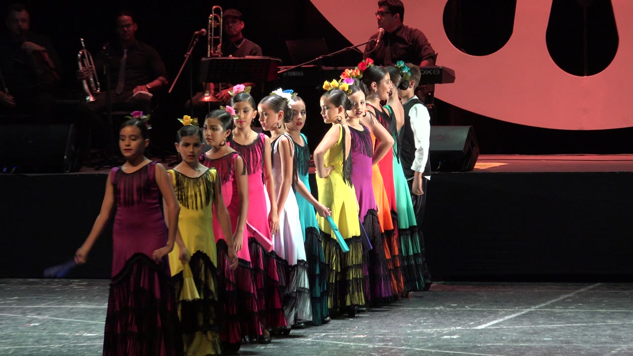 At 0:00 in Cabo San Lucas, young girls in bright dresses stand in a diagonal line on a stage while musicians are visible in the dark background.
