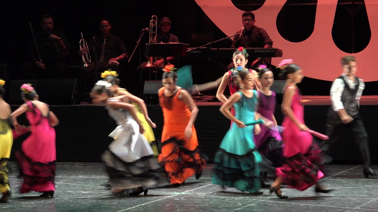 Girls in colorful dresses dance across a stage with musicians behind them.