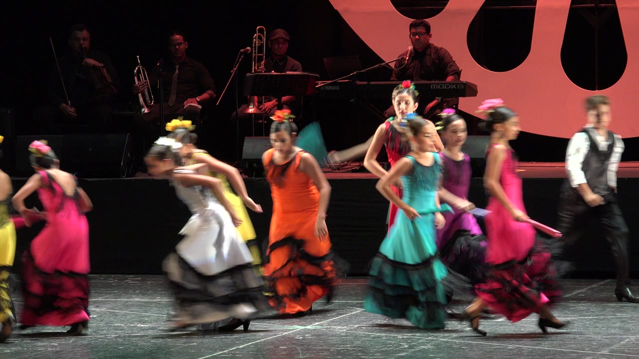 In Cabo San Lucas, Mexico, children in layered dresses pass one another onstage as a boy or young performer in black appears near the right edge and musicians sit behind them.