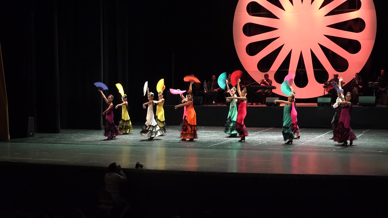 In Cabo San Lucas, Mexico, young dancers hold colorful fans in a line on a lit theater stage while musicians sit behind a large pink circular backdrop.