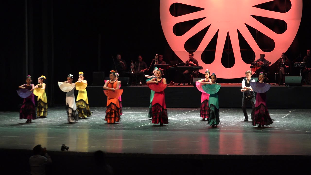 Women perform with fans on a stage in front of a red circular backdrop.
