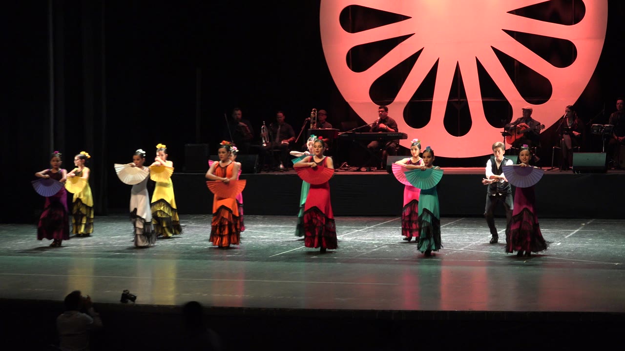 The dance line remains wide across the Cabo San Lucas stage, with women in red, teal, yellow, and purple dresses holding fans as the band sits in shadow behind them.