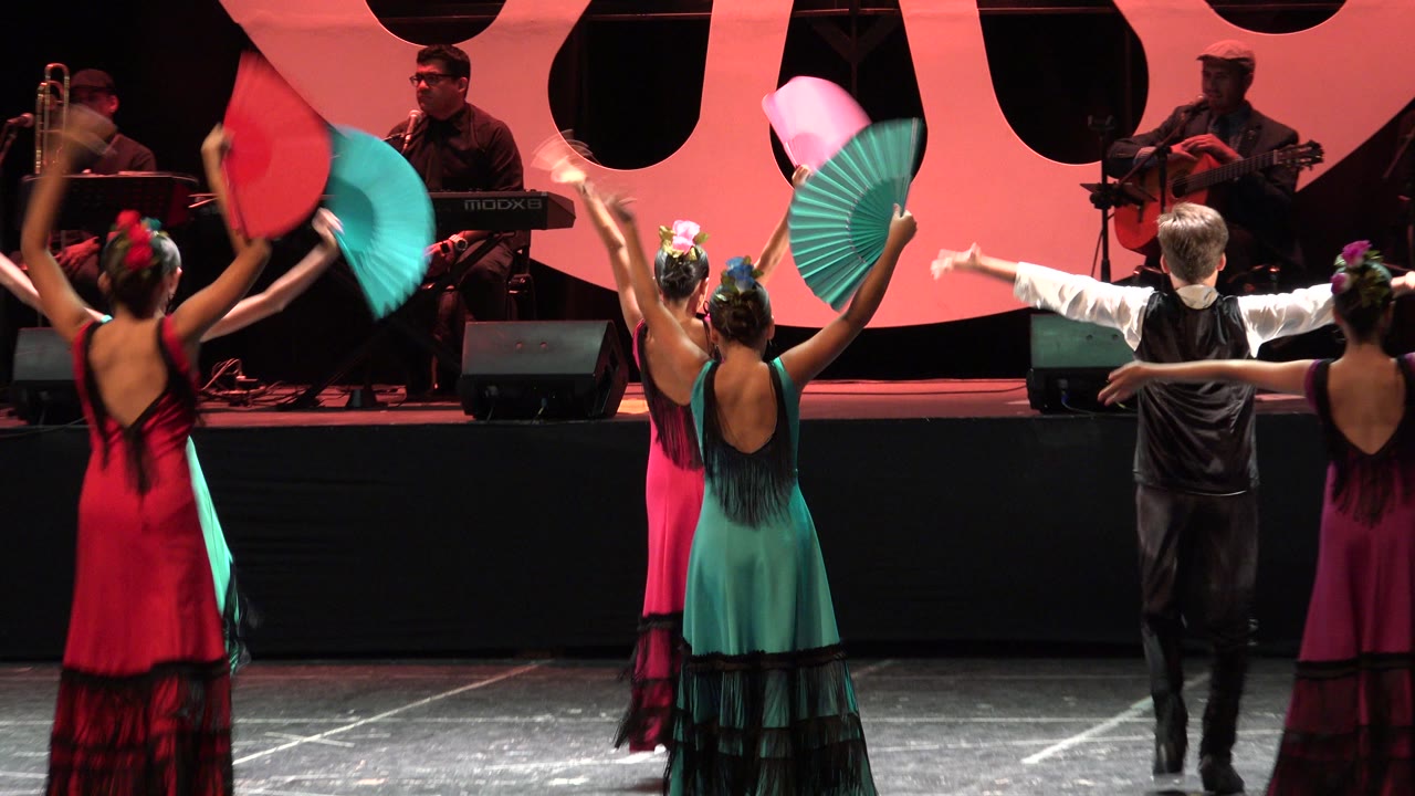 In Cabo San Lucas, Mexico, young dancers hold turquoise and pink fans aloft while a boy in a black vest stands center stage facing the musicians.