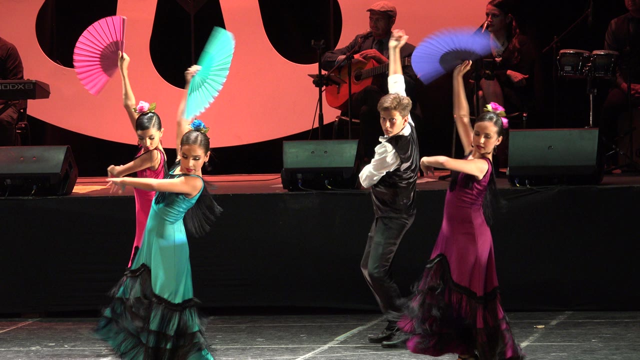 Children perform with turquoise, pink, and purple fans on a lit Cabo San Lucas stage while musicians sit in the darker background.