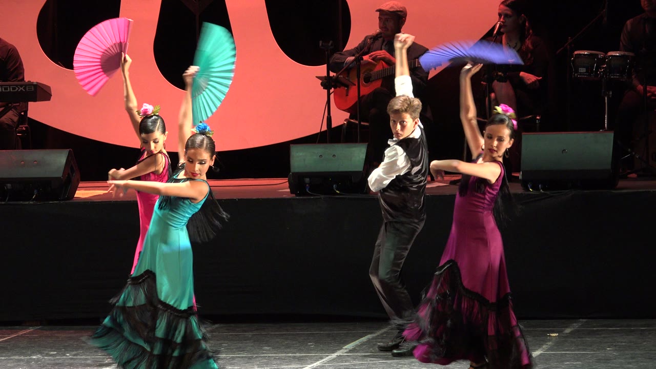 A boy in a black vest turns between two girls in bright dresses as raised fans cross the red-and-black stage backdrop in Cabo San Lucas.