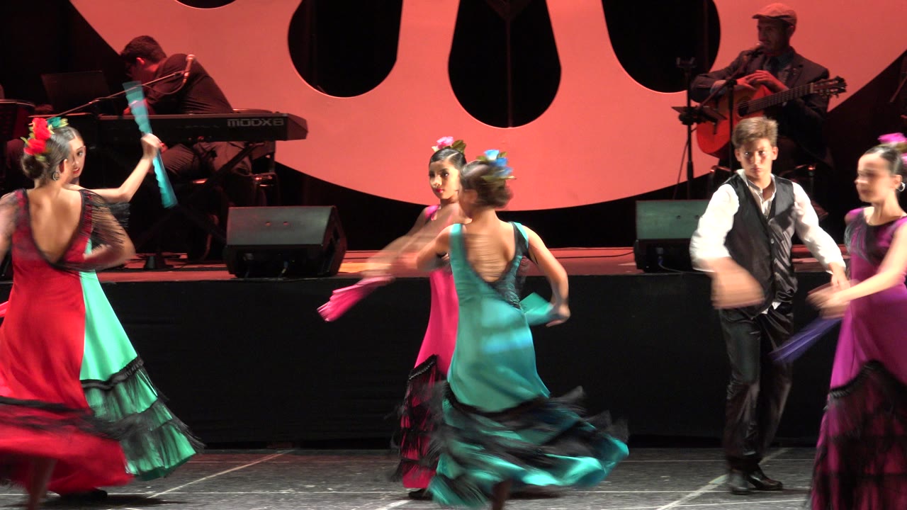 Teen dancers in red, turquoise, and pink ruffled dresses perform on a stage in Cabo San Lucas, Mexico, with musicians and microphones visible in the background.