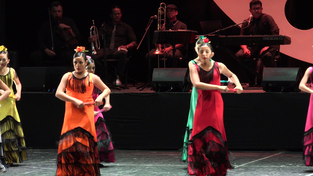 Young dancers in colorful dresses perform on a stage with musicians behind them.