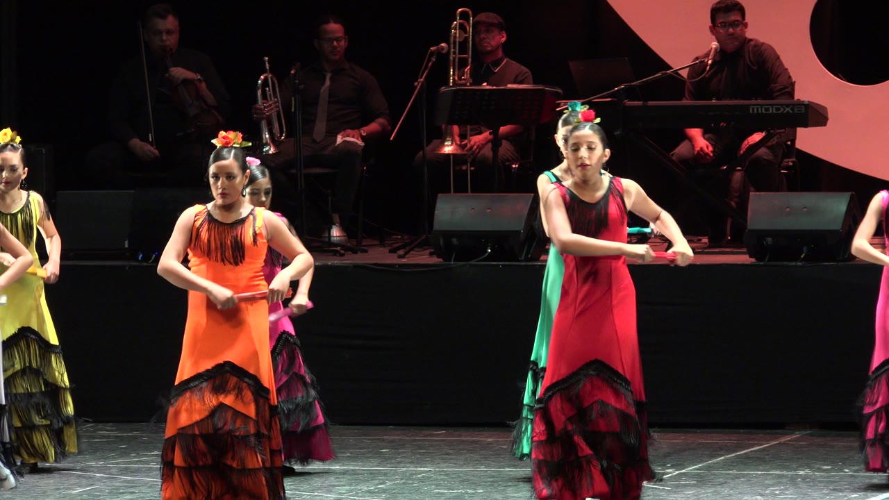 One second into the Cabo San Lucas performance, dancers in red and orange dresses hold a forward arm position as other young performers line up behind them near the musicians.