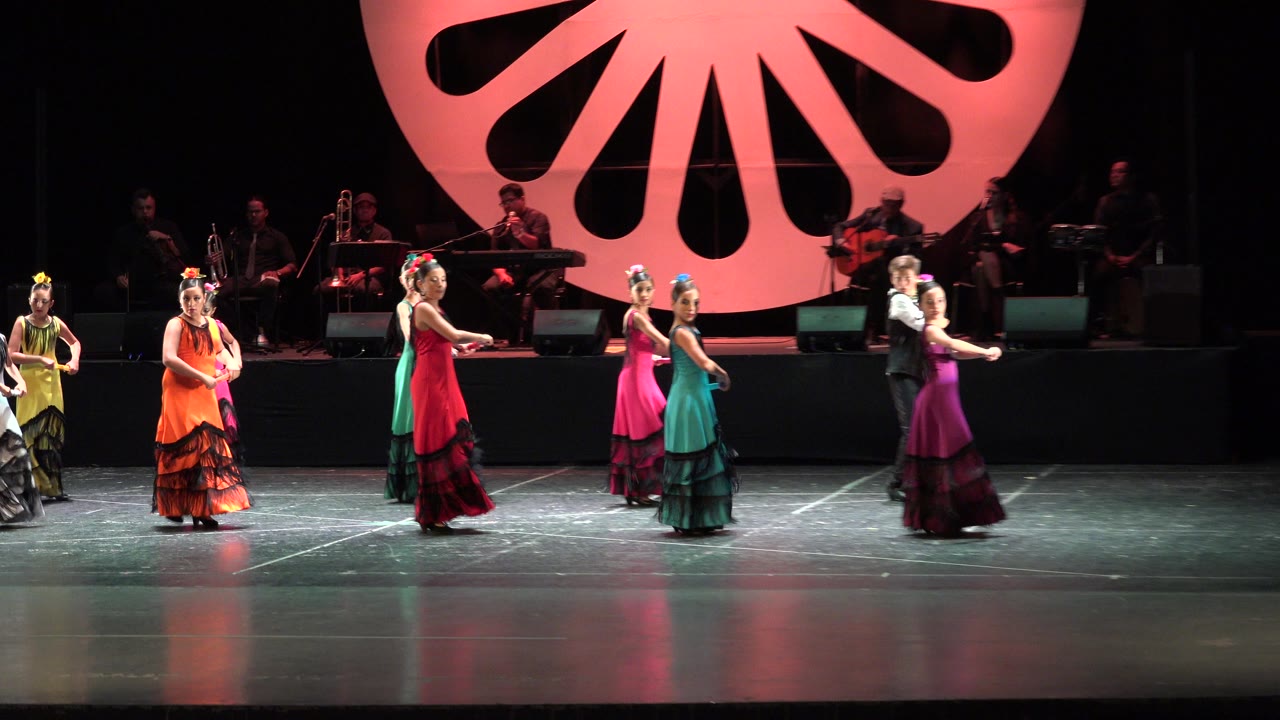 Women in bright dresses dancing on a dark stage with musicians behind them.