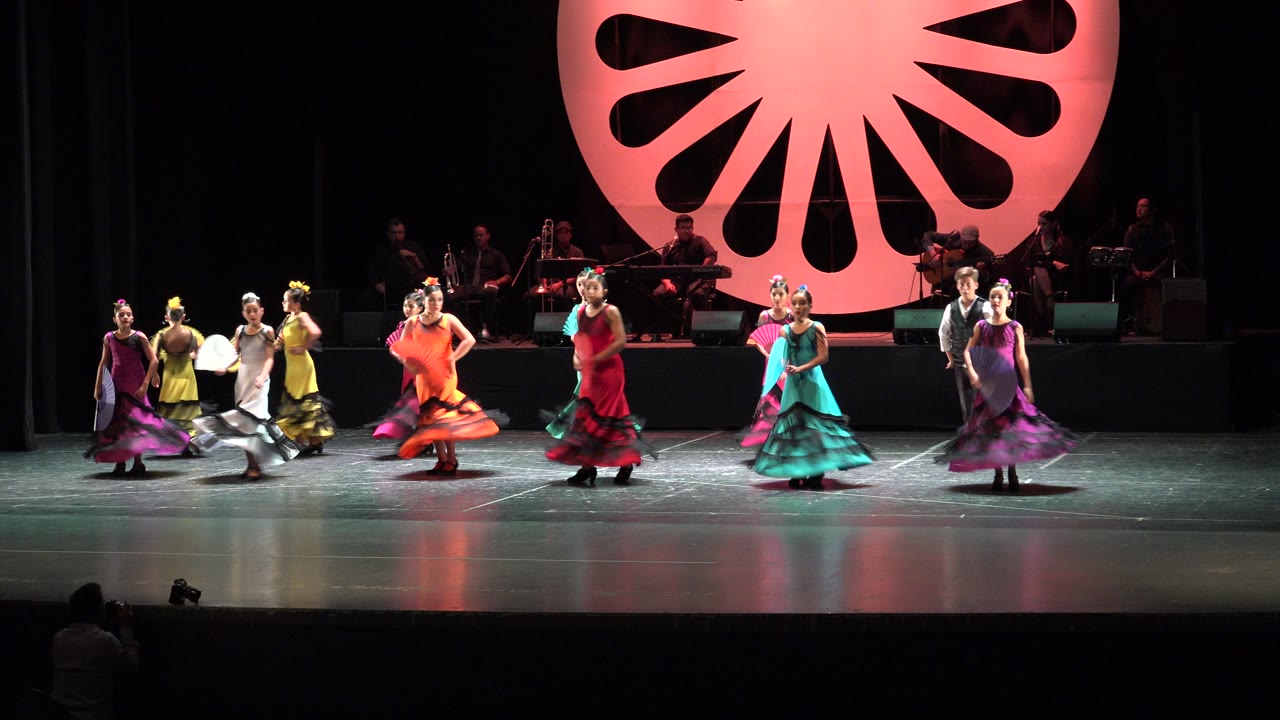 A wide stage moment in Cabo San Lucas, Mexico shows dancers in orange, red, teal, purple, yellow, and white costumes facing the audience under bright overhead light.