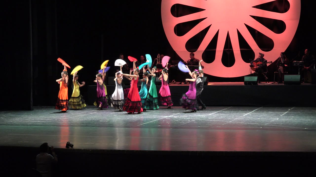 Colorful dancers perform on a theater stage with a large pink backdrop.