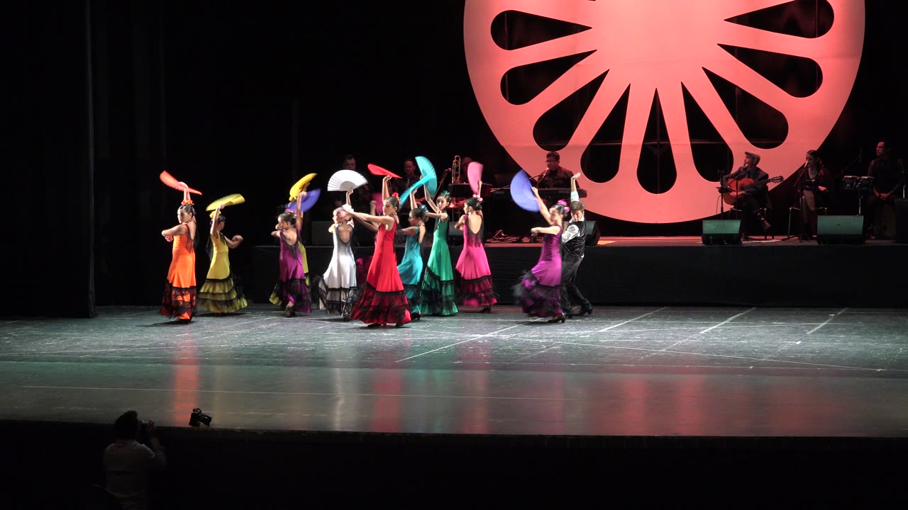 Dancers in orange, yellow, white, red, teal, and purple costumes hold fans aloft during a stage performance in Cabo San Lucas, Mexico.