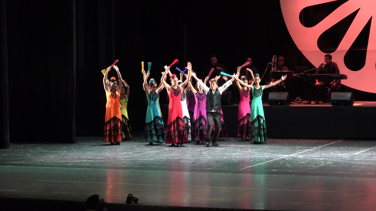 In Cabo San Lucas, Mexico, young performers in orange, teal, red, purple, black, and green costumes stand in a line with arms raised while musicians sit in the dark background.