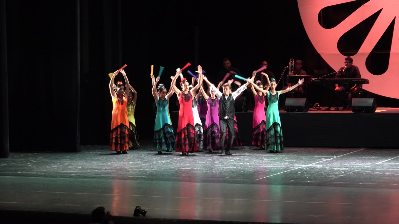 The dance group remains in a tight line on the Cabo San Lucas stage, with several performers lifting props or hands overhead and the band visible behind them at right.