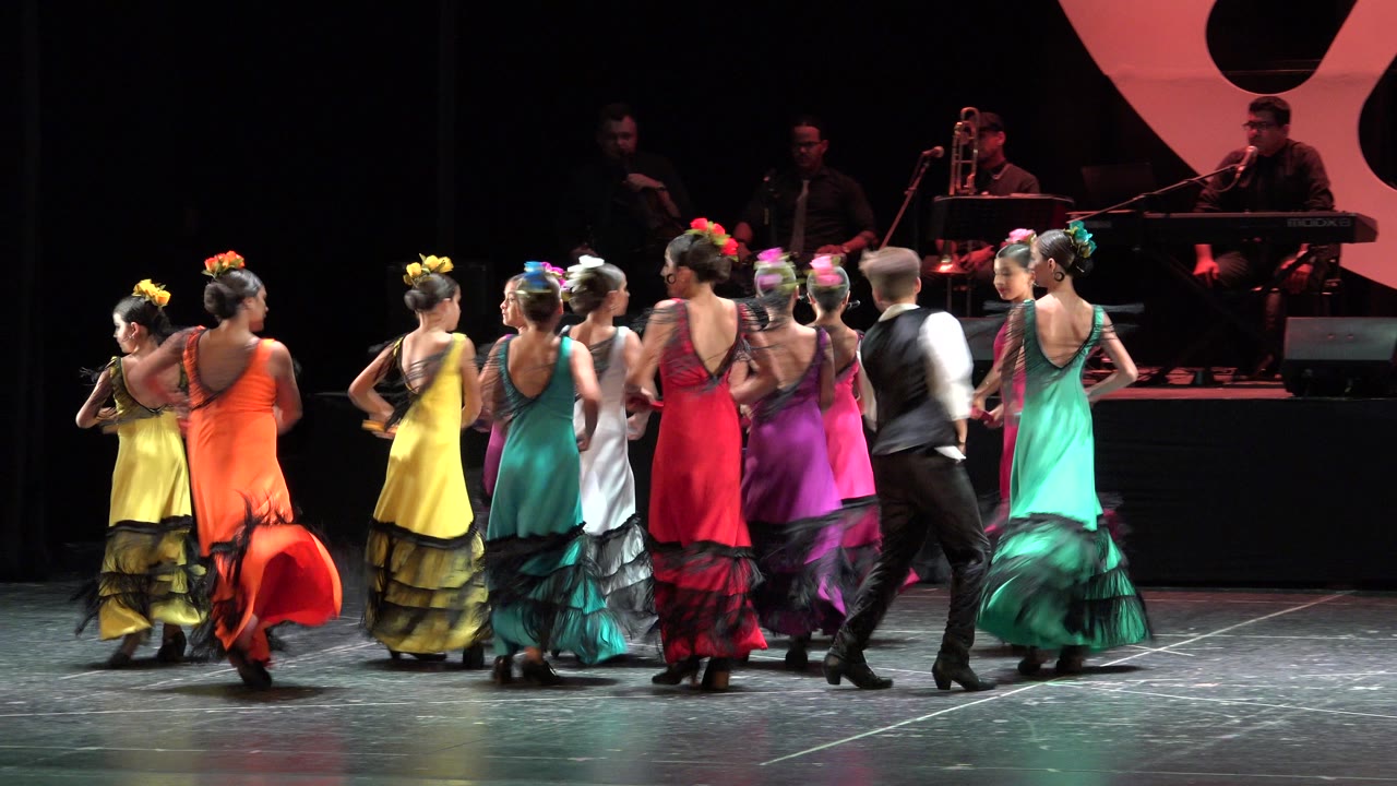 Dancers in colorful dresses move across a dark stage with musicians behind them.