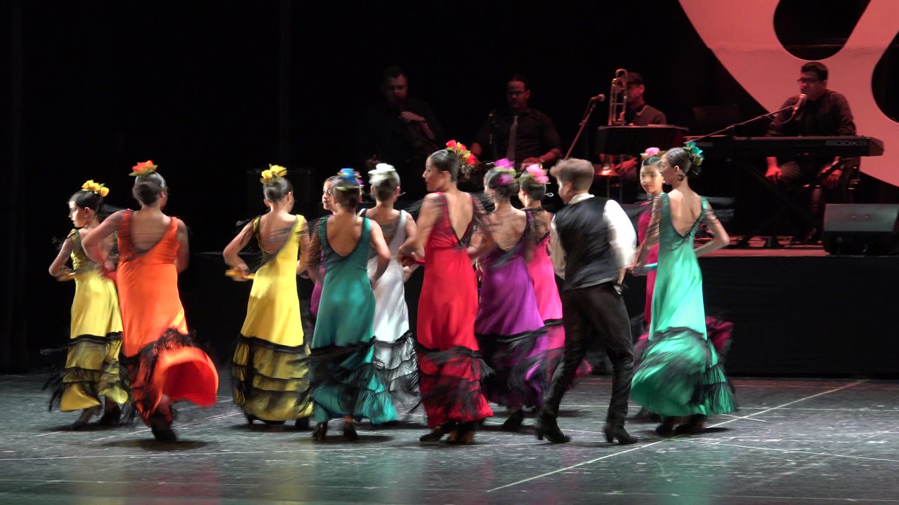 The ensemble continues the stage routine in Cabo San Lucas, Mexico, with dancers seen mostly from behind and musicians seated behind them.