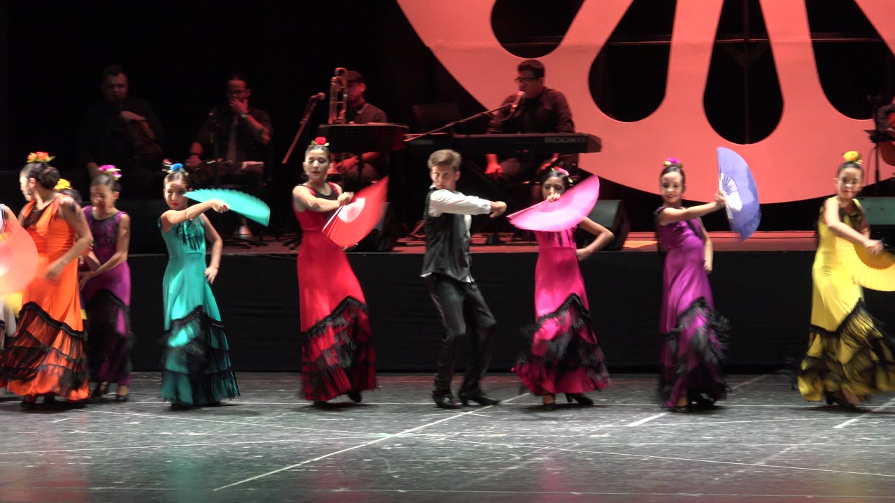 A boy in a black vest and dancers in red, pink, teal, yellow, and purple costumes perform with paper fans on stage in Cabo San Lucas.