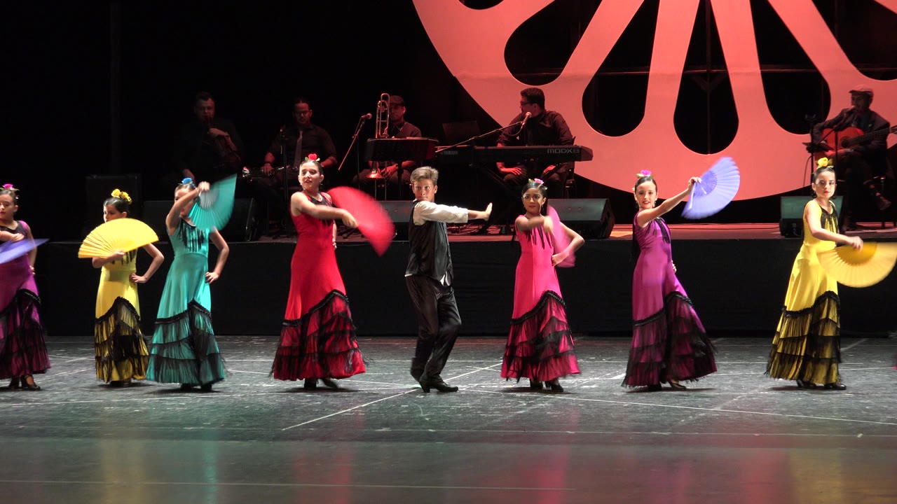 Wide view of a Cabo San Lucas stage performance with dancers in bright dresses and fans, a male performer near center, musicians in the dark background, and an orange fan-shaped backdrop.