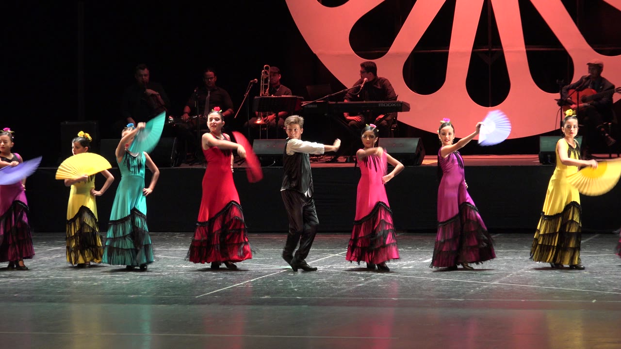 Dancers on the Cabo San Lucas stage hold yellow, teal, red, pink, and blue fans while the central male performer gestures forward under bright stage lights.