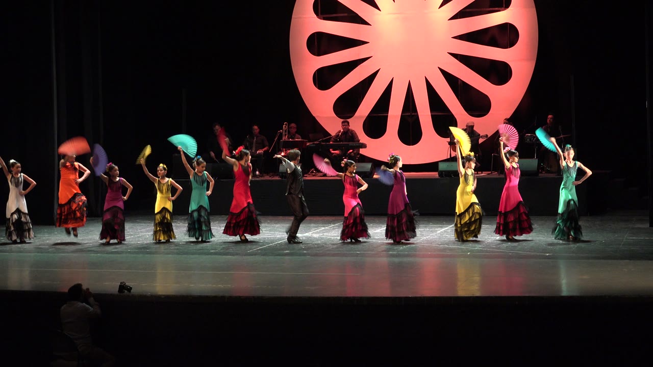 Wide theater stage with dancers, musicians, fans, and an orange circular backdrop.