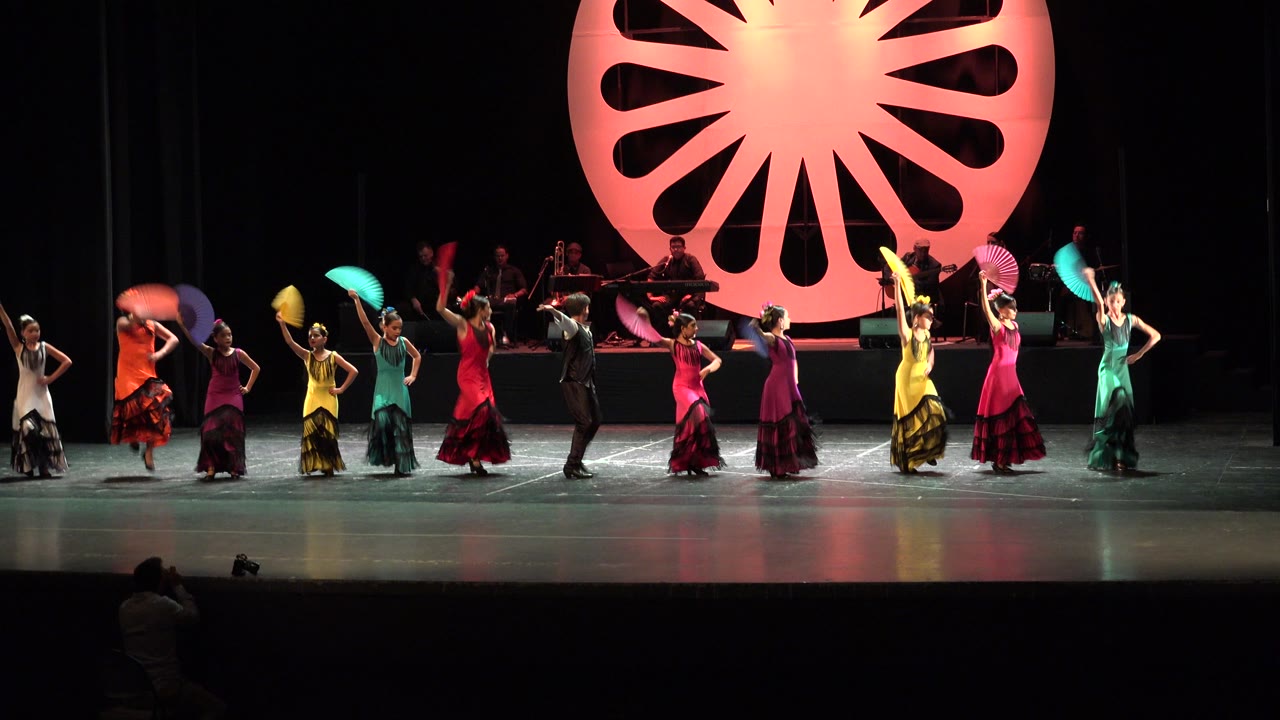 At 0:01 in Cabo San Lucas, the performer line shifts across the stage as several dancers lift turquoise, yellow, pink, and orange fans in front of the seated band.